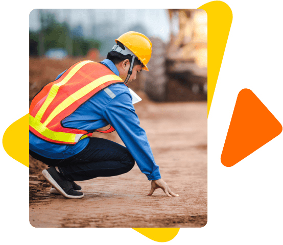 Homem em um canteiro de obras, de capacete amarelo e abaixado com a mão no chão. Ao fundo, desfocado, a imágem de uma grande máquina amarela.