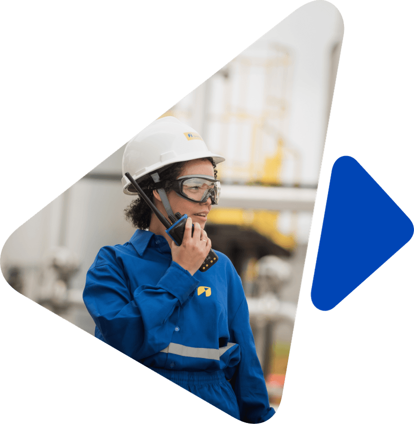 Mulher com cabelos pretos e cacheados, utilizando um capacete branco e uniforme azul, falando ao rádio. Ao fundo, uma imagem desfocada de uma indústria.